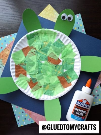 A Paper Plate Turtle craft made with green and brown tissue paper, green paper flippers and head, googly eyes, Elmer’s glue, and colorful papers in the background—a fun way to create your own adorable paper turtle!.