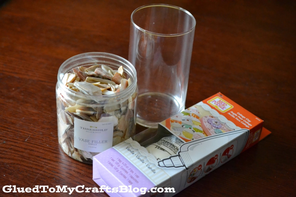 A clear glass vase, a jar of assorted seashells labeled vase filler, and a box of decorative white sand are arranged on a wooden surface—perfect for creating your own DIY shell vase.
