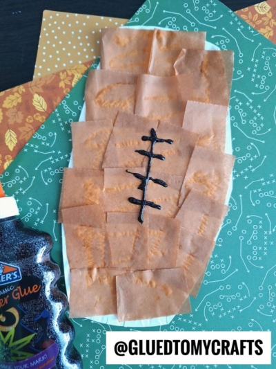 A paper plate football craft shaped like a foot, covered in brown paper squares with a black stitch pattern drawn on it. Orange glitter glue and patterned papers are nearby. @GLUEDTOMYCRAFTS is written at the bottom.