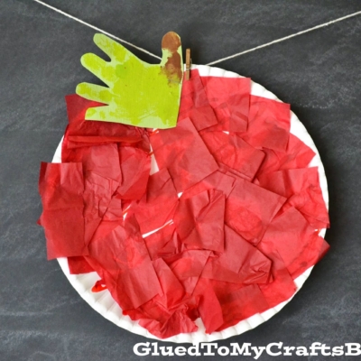 A tissue paper apple craft features a paper plate covered in red tissue, with a green handprint leaf. Hanging by a string and clothespin, this charming Tissue Paper Apple is showcased with GluedToMyCraftsBlog.com in the corner.