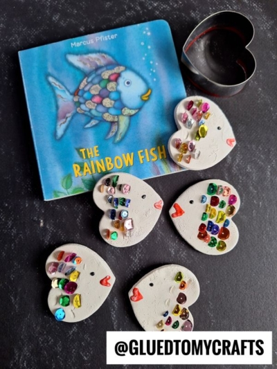 A children’s book titled The Rainbow Fish is shown with five heart-shaped clay rainbow fish decorated with colorful gems and red lips, plus a metal heart-shaped cookie cutter. The handle @GLUEDTOMYCRAFTS is displayed.