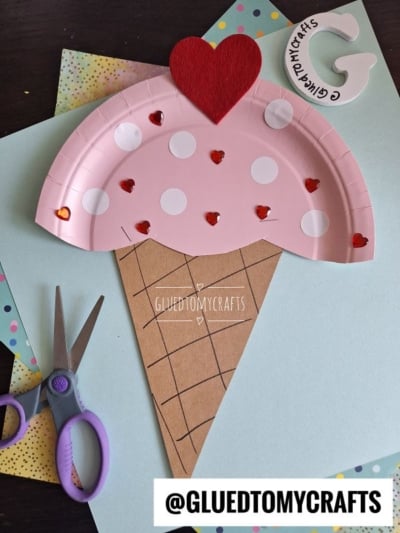 A paper plate ice cream kid craft made from a pink polka dot plate with heart-shaped decorations on a brown cone. Scissors and pastel papers are nearby. Text reads @GLUEDTOMYCRAFTS.