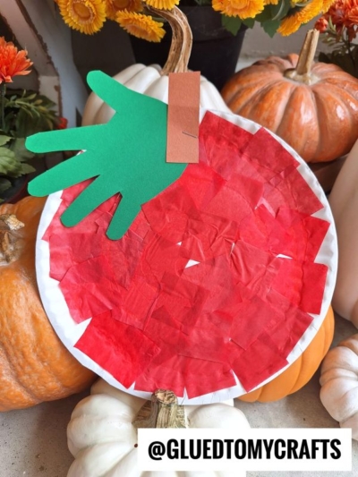 A paper plate apple craft with red tissue paper, a green paper handprint leaf, and a brown stem, placed among pumpkins and autumn flowers. Perfect for a fun tissue paper apple tree craft! Text: @GLUEDTOMYCRAFTS.