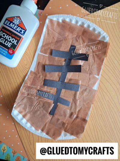 A paper plate football craft is displayed, decorated with brown tissue, black paper strips shaped like football laces, and a bottle of Elmer’s glue beside it. Craft supplies are arranged on a patterned surface.