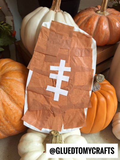 A paper plate football made with brown and white paper sits among orange and white pumpkins. The football features a white lacing pattern and is displayed upright. The text @GLUEDTOMYCRAFTS is at the bottom.