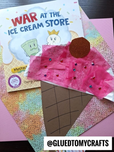 A children’s book titled War at the Ice Cream Store lies on colorful paper. On top is a paper plate ice cream kid craft—an ice cream cone with a pink scoop, brown cone, red cherry, and tiny beads. @GLUEDTOMYCRAFTS is visible.