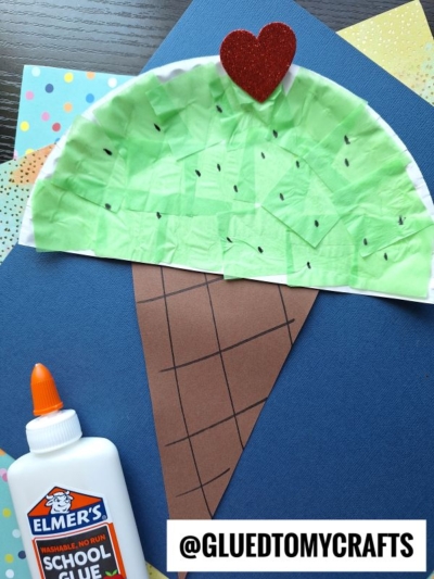 A fun ice cream kid craft: a watermelon slice made with green tissue paper, a brown paper cone, black marker seeds, red glitter heart, and Elmers glue. Perfect for summer! @GLUEDTOMYCRAFTS is written at the bottom.