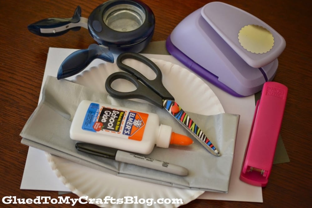 Craft supplies arranged on a table, including scissors, glue, stapler, Sharpie, paper plates—perfect for making a Paper Plate Shark—plus tissue paper, craft punches, and paper. GluedToMyCraftsBlog.com text at the bottom.