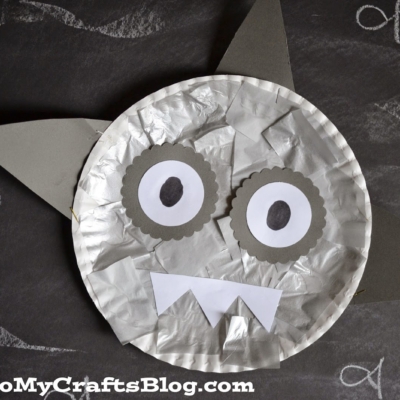 A Paper Plate Shark craft featuring gray fins, big round eyes, and triangle teeth is displayed on a chalkboard background with white chalk swirls.
