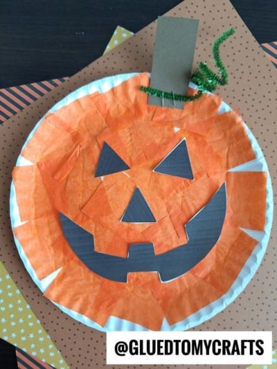 Paper plate pumpkin craft decorated with orange tissue paper, black paper facial features, a green pipe cleaner stem, and a brown paper rectangle stem, sitting on patterned orange and yellow paper. This easy and fun project is perfect if you’re looking for how to make a pumpkin with a paper plate. @GLUEDTOMYCRAFTS is printed at the bottom.
