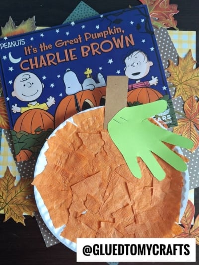 A children’s Paper Plate Pumpkin craft made with orange tissue on a paper plate and a green handprint leaf, placed on fall leaves alongside the book “It’s the Great Pumpkin, Charlie Brown.” Perfect for showing how to make a pumpkin with a paper plate. @GLUEDTOMYCRAFTS is written at the bottom.