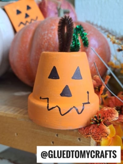An orange mini flowerpot painted as a jack-o-lantern with a brown pipe cleaner stem and green leaf, perfectly capturing the charm of a Terra Cotta Pot Pumpkin. A pumpkin and fall flowers are in the background. Text at the bottom reads @GLUEDTOMYCRAFTS.