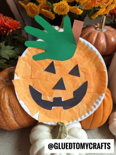 A paper plate pumpkin craft featuring vibrant orange tissue paper, a black cut-out face, and a green handprint stem, surrounded by pumpkins and flowers. This charming scene perfectly captures how to make a pumpkin with a paper plate—simple, fun, and festive! @GLUETOMYCRAFTS text at the bottom.