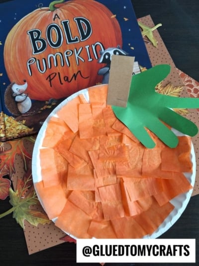 A pumpkin-themed kids craft made with a paper plate, orange tissue paper, and a green handprint stem—showing how to make a pumpkin with a paper plate—set on a book called A Bold Pumpkin Plan and autumn-themed paper. @gluedtomycrafts.