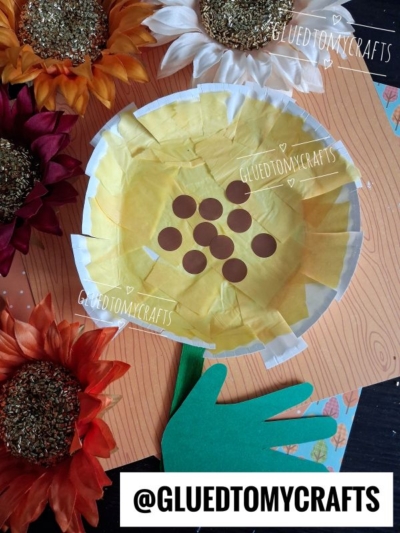 A handmade paper plate sunflower with a green hand-shaped stem, surrounded by artificial sunflowers, sits on a wood-patterned background. The image is watermarked with @GLUEDTOMYCRAFTS.