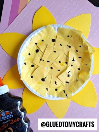 A paper plate sunflower made from yellow paper petals and tissue squares on a white plate, with black glitter glue details, sits on layered pink and purple paper. @GLUEDTOMYCRAFTS is visible at the bottom.