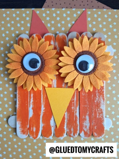 Craft a charming popsicle stick owl using orange and white-painted sticks, sunflower-shaped googly eyes, orange triangle ears, and a yellow beak—all set on a star-patterned background. @GLUEDTOMYCRAFTS at the bottom. #PopsicleStickOwls.