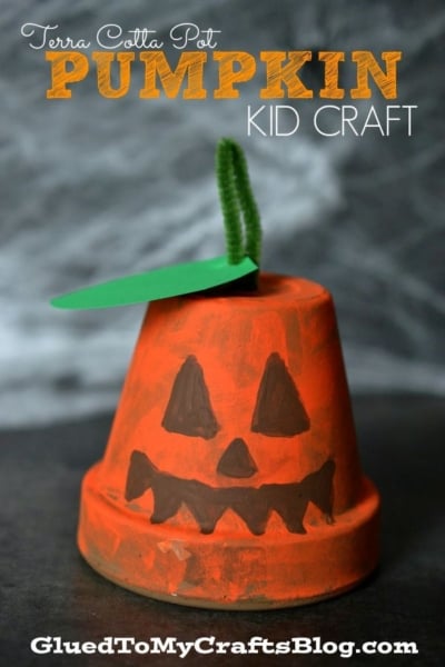 A small terra cotta pot painted orange to look like a Terra Cotta Pot Pumpkin, complete with a jack-o-lantern face, a green paper leaf, and a green pipe cleaner stem on top. Text reads: Terra Cotta Pot Pumpkin Kid Craft.