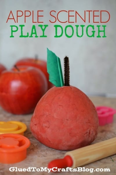 Red apple-scented play dough shaped like an apple, with a brown stem and green leaf. Play dough tools and red apples are in the background. Text reads Apple Scented Play Dough—perfect for creative fun with this delightful apple playdough!.