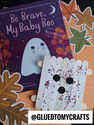 A book titled Be Brave, My Baby Boo features a cute ghost, autumn leaves, and a Popsicle Stick Ghost craft decorated with confetti and black circles, all beautifully arranged on an orange polka-dot background.