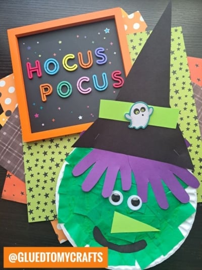 Halloween craft scene with HOCUS POCUS in colorful letters in a frame, and a paper plate halloween witch face with a black hat, googly eyes, and green tissue paper, all arranged on patterned orange, green, and brown papers.