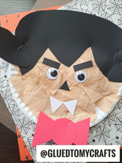 A Halloween craft of a Paper Plate Vampire, featuring tissue paper, large googly eyes, black paper hair and eyebrows, and a red paper bow tie, displayed on a spiderweb-patterned background.