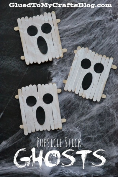 Three popsicle stick ghost faces made from white popsicle sticks with black oval eyes and mouths, arranged on a dark surface with fake spider webs. Text reads: Popsicle Stick Ghosts GluedToMyCraftsBlog.com.