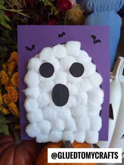 A ghost cotton ball craft is glued to purple paper with black paper eyes and mouth, plus bat cutouts. Flowers and pumpkins are in the background. Text reads @GLUEDTOMYCRAFTS.