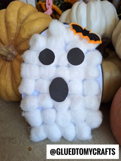 A ghost cotton ball craft, made with a cotton ball ghost template and black paper eyes and mouth, is topped with a small orange-and-black bat decoration and placed among pumpkins. The @GLUEDTOMYCRAFTS handle appears at the bottom.