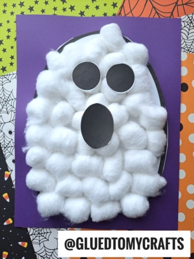 A cotton ball ghost with black paper eyes and mouth sits on a purple background. Using a cotton ball ghost template for this ghost cotton ball craft, Halloween-themed paper with spiderwebs and candy corn patterns surrounds it.