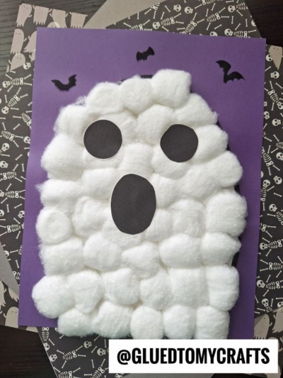 A halloween cotton ball ghost with black paper eyes and mouth sits on a purple background, surrounded by bat cutouts and skeleton-patterned paper. @GLUEDTOMYCRAFTS is written at the bottom.