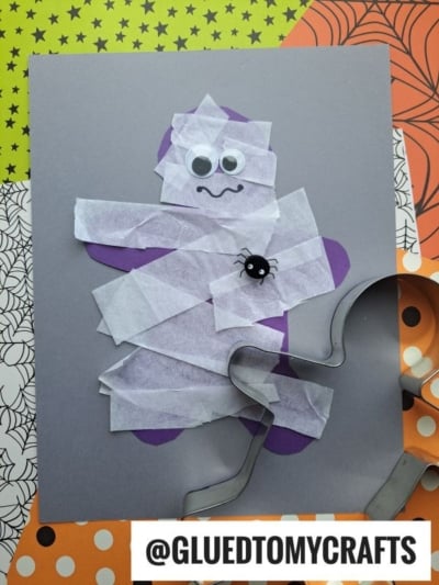 A Tissue Paper Mummy craft made with white tissue strips, googly eyes, and a small spider decoration on gray paper. A gingerbread man cookie cutter is nearby, with Halloween-themed backgrounds. Text: @GLUEDTOMYCRAFTS.