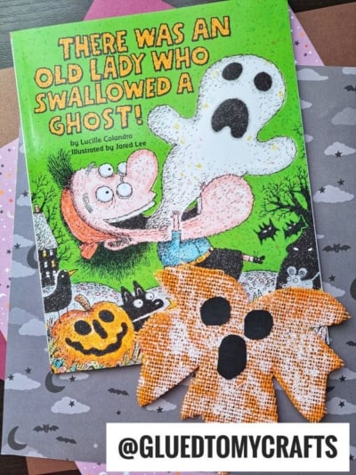 A children’s book titled “There Was an Old Lady Who Swallowed a Ghost!” lies on star and cloud-patterned paper, with two orange doily ghosts, painted ghost leaves, and a pumpkin craft nearby. Text at the bottom reads @GLUEDTOMYCRAFTS.