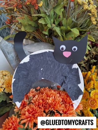 A Paper Plate Black Cat with googly eyes and a pink nose sits among orange and yellow flowers. Text at the bottom reads @GLUEDTOMYCRAFTS.