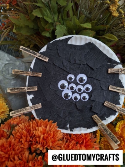 A Paper Plate Halloween Spider craft made with black crepe paper, multiple googly eyes, and clothespin legs, surrounded by flowers. Text: @GLUEDTOMYCRAFTS.