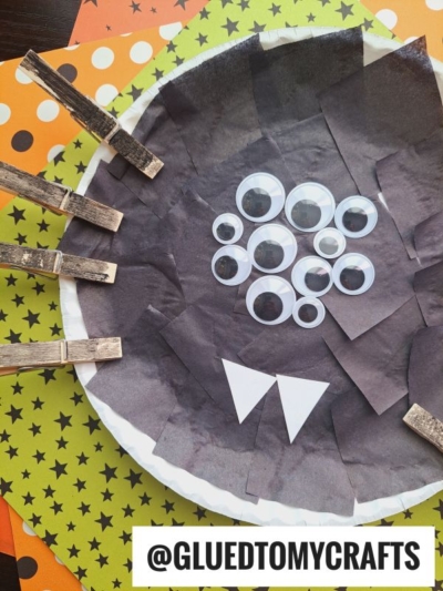 A Paper Plate Halloween Spider craft made from a paper plate with black tissue, googly eyes, clothespin arms, and white paper teeth. The plate sits on colorful star and polka dot paper. Text: @GLUEDTOMYCRAFTS.