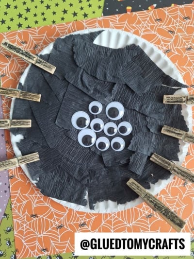 A Paper Plate Halloween Spider is crafted by covering a plate with strips of black paper and adding various sizes of googly eyes in the center. Clothespins serve as legs, and the spider sits on Halloween-themed papers with spiderwebs and stars.