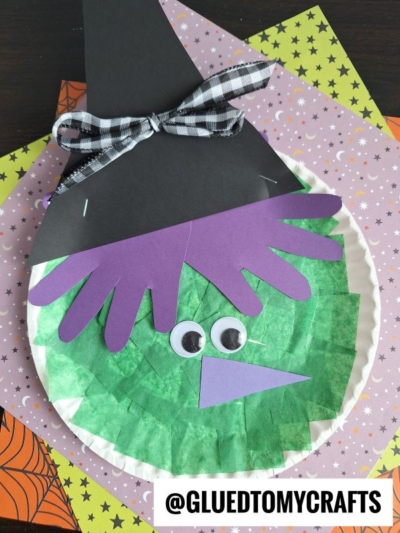 A paper plate halloween witch craft with green tissue paper, googly eyes, a purple nose, and purple paper hair. It features a black paper hat with a plaid ribbon, all displayed on colorful Halloween-themed paper.