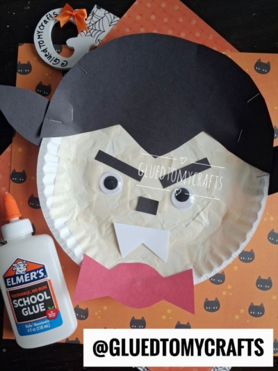 A paper plate Dracula craft featuring a vampire face with fangs, googly eyes, and black paper hair sits alongside a bottle of Elmer’s glue on a pumpkin and bat-themed background. @GLUEDTOMYCRAFTS is written on the image. This charming Paper Plate Vampire project is perfect for spooky season crafting!.