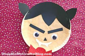 A Paper Plate Vampire craft featuring black hair, pointed ears, googly eyes, fangs, and a red tongue on a red background with white dots. Perfect paper plate dracula project! GluedtoMyCraftsBlog.com is at the bottom.