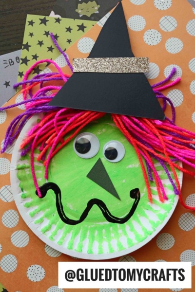 A paper plate Halloween witch with googly eyes, yarn hair, a black hat, and a drawn-on smile sits on patterned paper. @GLUEDTOMYCRAFTS text is displayed at the bottom.
