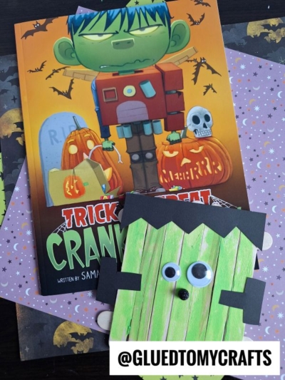 A Halloween-themed children’s book titled “Trick or Treat Crankenstein” is displayed with a green, Popsicle Stick Frankenstein craft featuring googly eyes, set on festive paper. A watermark reads “@GLUEDTOMYCRAFTS.”.