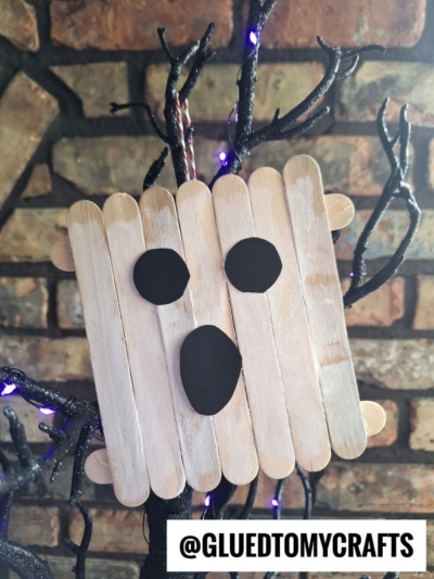 A spooky Popsicle Stick Ghost face made from craft sticks and black paper shapes is displayed on black, branch-like decorations with purple lights, against a brick wall. The text @GLUETOMYCRAFTS appears at the bottom.