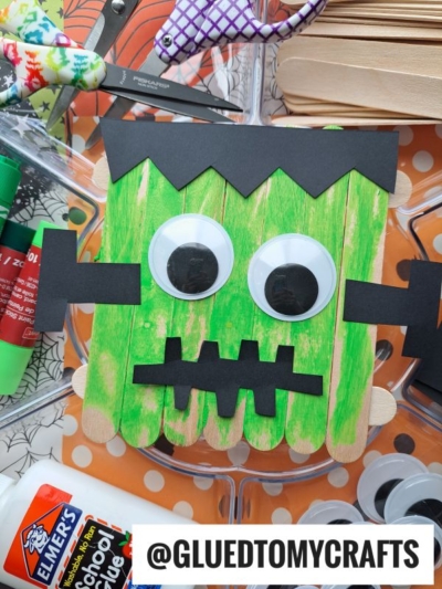 A Popsicle Stick Frankenstein monster craft made from green-painted popsicle sticks, large googly eyes, and black paper features is surrounded by craft supplies like glue, markers, and foam shapes.