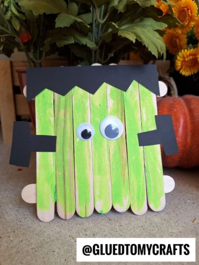 A Popsicle Stick Frankenstein monster craft made from green-painted sticks, black paper hair and bolts, and two large googly eyes. Autumn flowers and a pumpkin are in the background. @GLUEDTOMYCRAFTS is at the bottom.