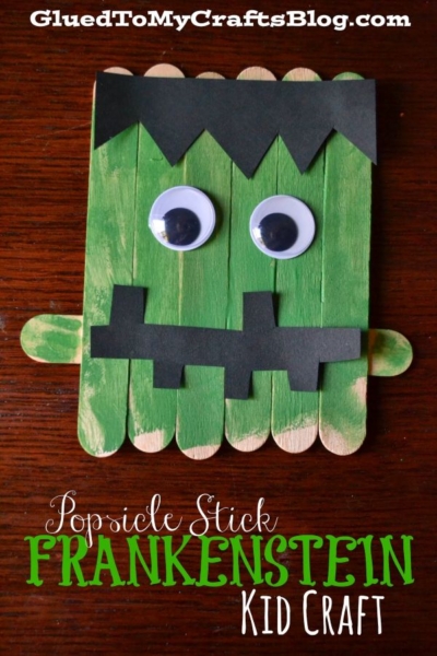 Green popsicle sticks are creatively arranged as a Popsicle Stick Frankenstein’s face, complete with large googly eyes, black paper hair, and a quirky mouth, all set on a dark wooden surface. Text: “Popsicle Stick Frankenstein Kid Craft.”.