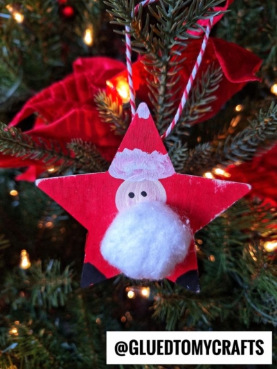A handmade Santa Star Ornament shaped like a red star with a fluffy white cotton ball beard hangs on a Christmas tree branch. In the background are green needles, lights, and a red bow. Text at the bottom reads @GLUEDTOMYCRAFTS.