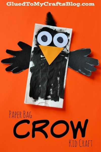 A charming paper bag decorated as a crow, featuring black wings, large eyes, and an orange beak, sits against an orange background. The words "Paper Bag Crow Kid Craft" are written at the bottom. This Paper Bag Crow makes a fun and creative project for kids to enjoy!.