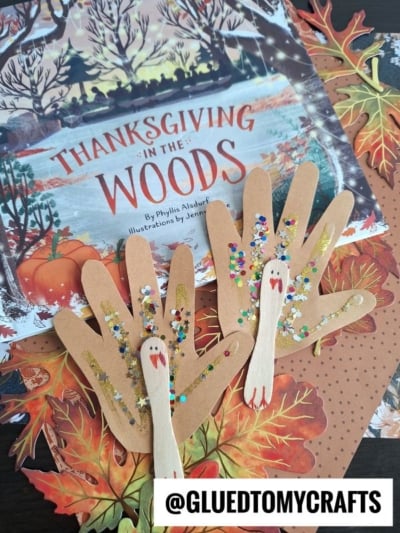 Two handprint turkeys with googly eyes and sequins are displayed on fall leaves beside a children’s book titled Thanksgiving in the Woods. A tag reads @GLUEDTOMYCRAFTS. These adorable Handprint Turkey Puppets make a festive Thanksgiving craft!.