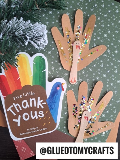 A card shaped like a turkey, reading Five Little Thank-yous, sits next to two sparkling Handprint Turkey Puppets with glitter, on a green patterned surface accented by pine branches. @GLUEDTOMYCRAFTS is written at the bottom.